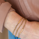 Three delicate gold chain bracelets with small letter charms worn stacked on a person's wrist, shown against a dusty rose top and blue denim background.