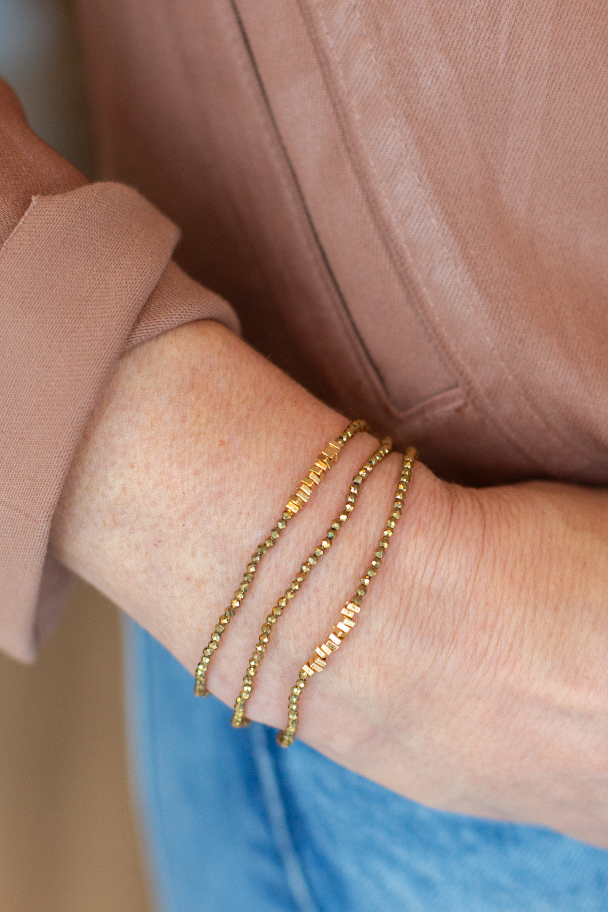 Three delicate gold chain bracelets with small letter charms worn stacked on a person's wrist, shown against a dusty rose top and blue denim background.