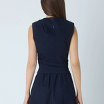 A woman viewed from the back wearing a sleeveless dark navy midi dress with a gathered waist and a small gold zipper detail at the neckline, set against a plain light gray studio background.