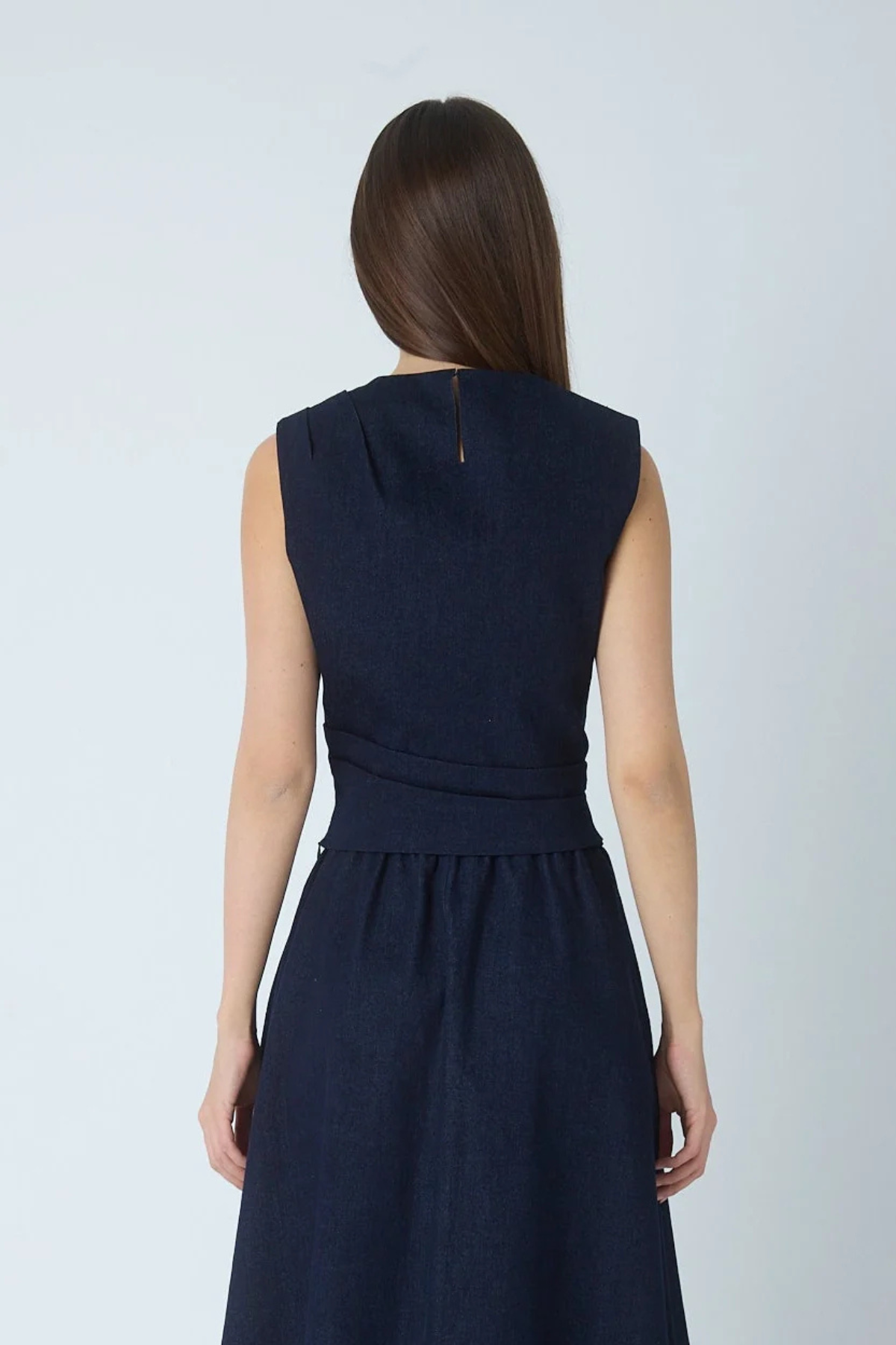 A woman viewed from the back wearing a sleeveless dark navy midi dress with a gathered waist and a small gold zipper detail at the neckline, set against a plain light gray studio background.