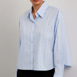 A woman wearing a light blue long-sleeve button-up shirt with a subtle gold geometric pattern, paired with black trousers, against a neutral gray studio background.