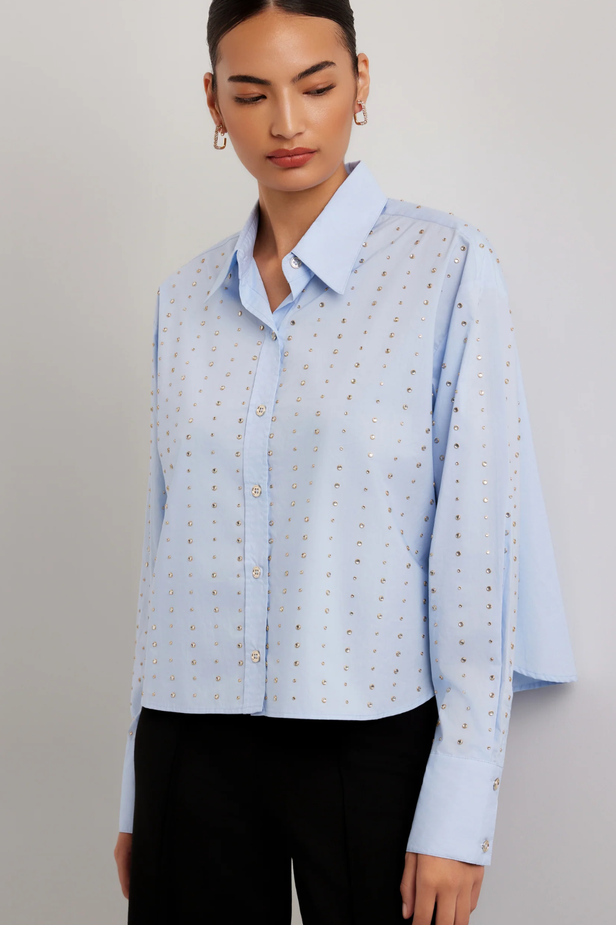 A woman wearing a light blue long-sleeve button-up shirt with a subtle gold geometric pattern, paired with black trousers, against a neutral gray studio background.
