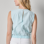 Woman wearing a light blue sleeveless top and white pants on a gray background