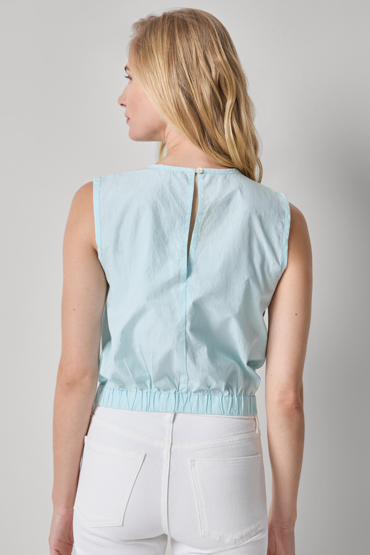 Woman wearing a light blue sleeveless top and white pants on a gray background