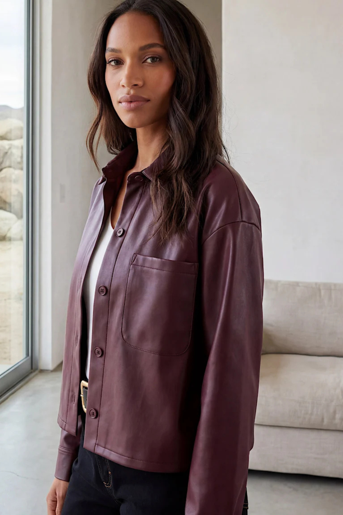 A woman wearing a cropped, long-sleeve button-up shirt jacket in a deep plum purple leather or faux-leather material, styled over a white top and black pants in a modern indoor setting.
