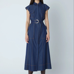 A woman modeling a dark blue denim midi dress with a matching belt and silver buckle, styled with brown leather boots against a light gray studio background.