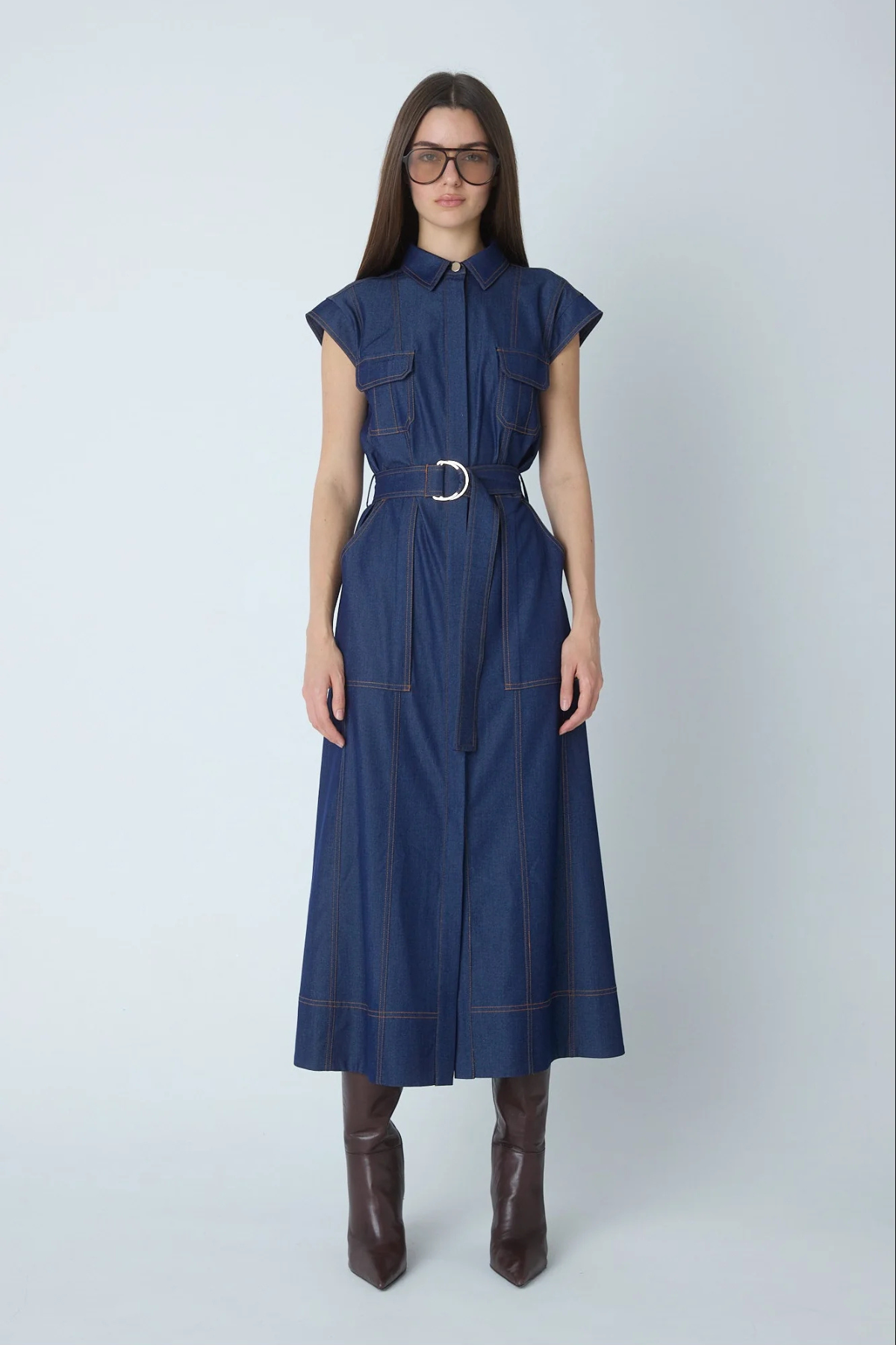 A woman modeling a dark blue denim midi dress with a matching belt and silver buckle, styled with brown leather boots against a light gray studio background.