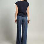 A woman shown from the back wearing a navy blue sleeveless top and wide-leg blue jeans, standing against a light gray studio background.