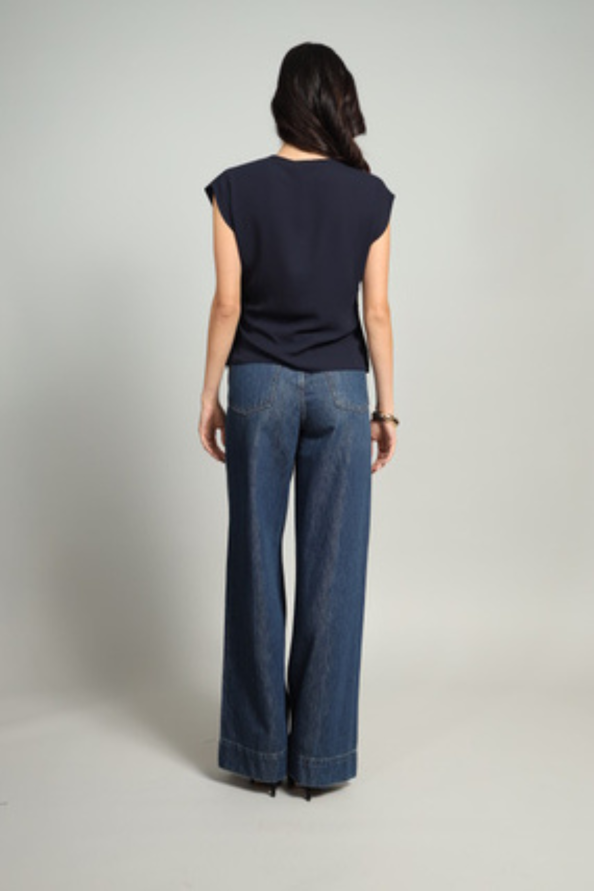 A woman shown from the back wearing a navy blue sleeveless top and wide-leg blue jeans, standing against a light gray studio background.