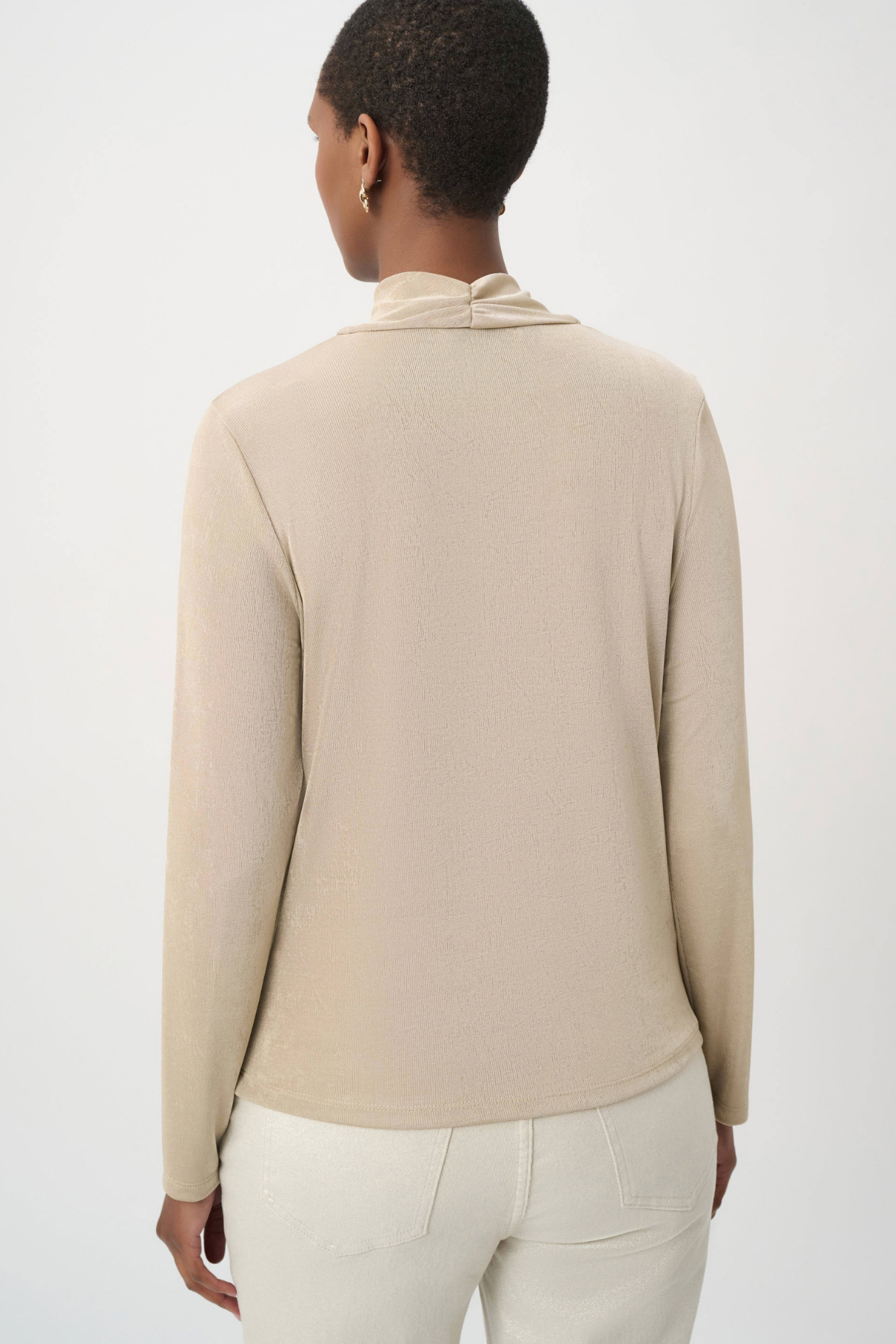 A woman shown from the back wearing a light beige long-sleeve knit top with a high folded collar, paired with off-white pants, against a plain light gray studio background.