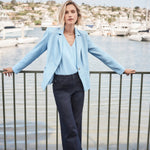Woman in light blue blazer and dark pants standing by a waterfront with boats in the background