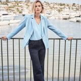 Woman in light blue blazer and dark pants standing by a waterfront with boats in the background