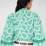 Woman wearing a green patterned blouse with white pants on a plain background
