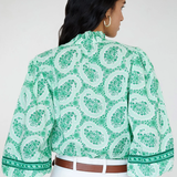 Woman wearing a green patterned blouse with white pants on a plain background