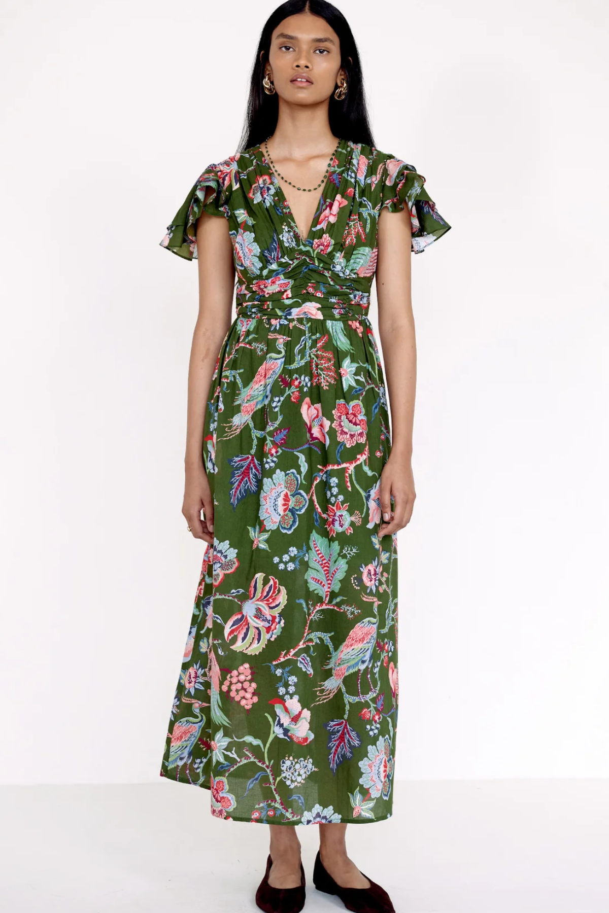 A model wearing a green midi dress with a colorful floral and bird print, featuring flutter sleeves and a tie-waist detail, set against a plain white studio background.