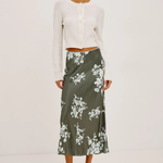 A model stands against a plain white studio background wearing a white cropped long-sleeve cardigan and a dark olive green midi skirt with a white floral print, paired with black strappy high-heeled sandals.