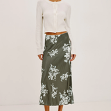 A model stands against a plain white studio background wearing a white cropped long-sleeve cardigan and a dark olive green midi skirt with a white floral print, paired with black strappy high-heeled sandals.