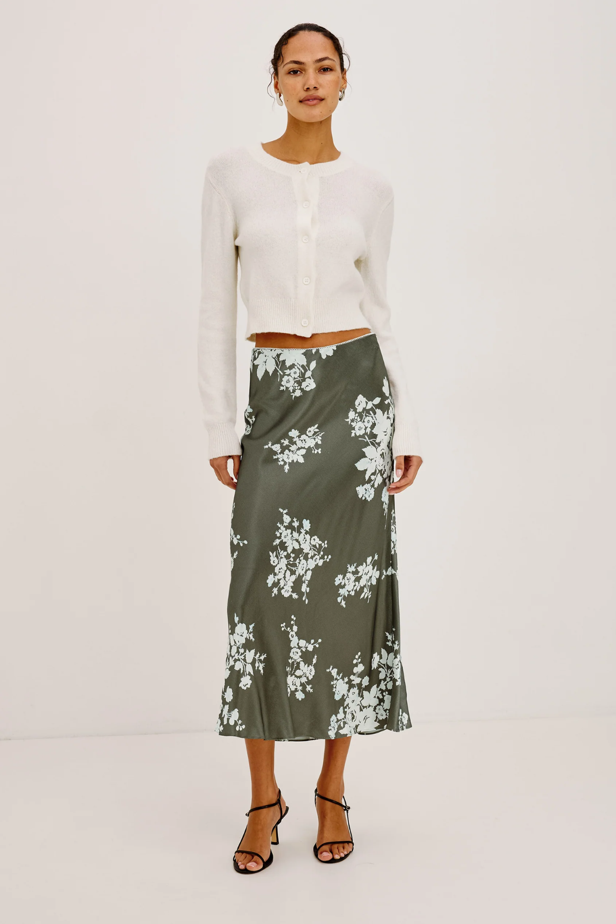 A model stands against a plain white studio background wearing a white cropped long-sleeve cardigan and a dark olive green midi skirt with a white floral print, paired with black strappy high-heeled sandals.