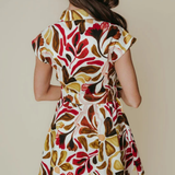 A woman shown from behind wearing a short-sleeve collared dress with a bold abstract floral pattern in red, brown, olive green, and cream on a light background.