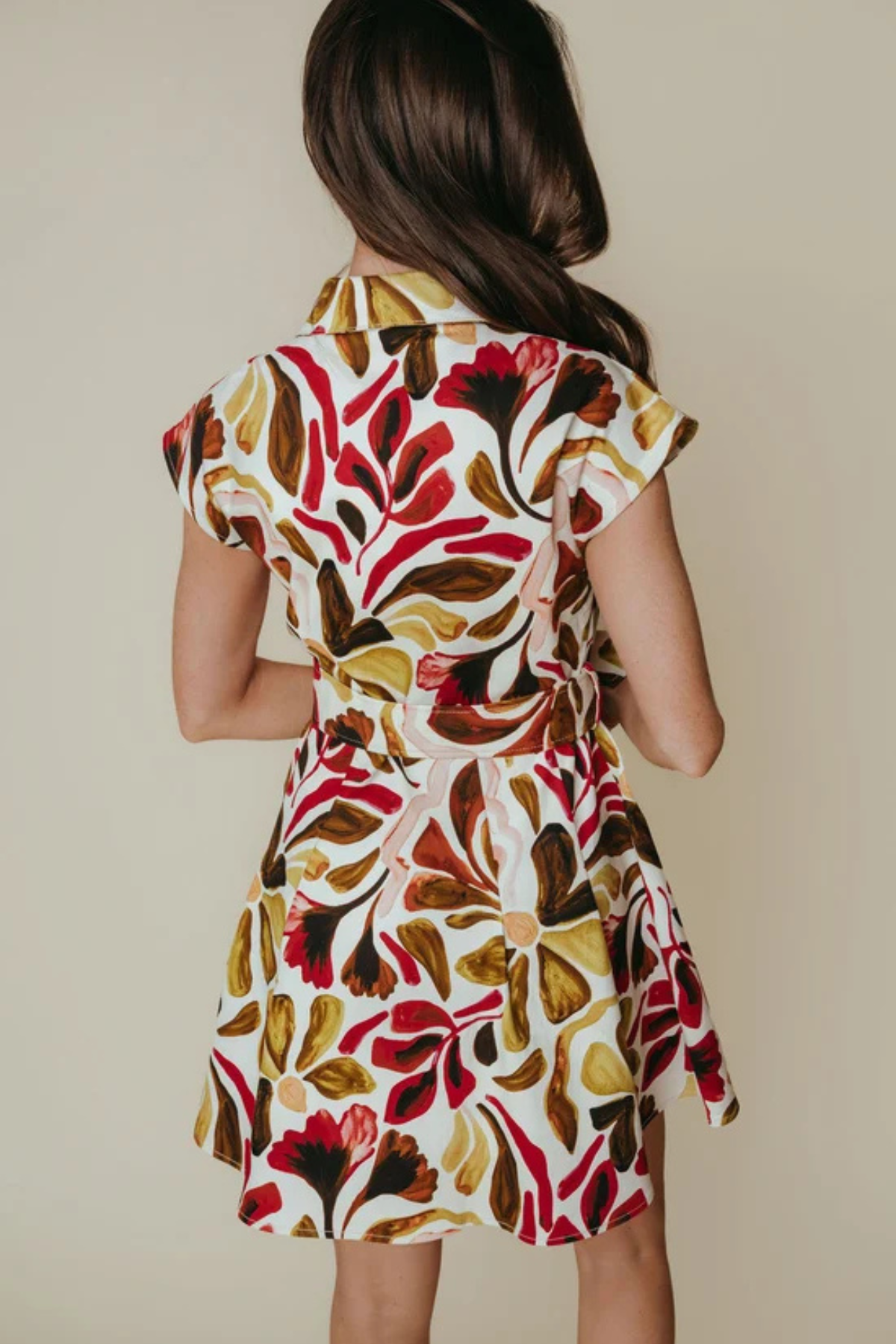 A woman shown from behind wearing a short-sleeve collared dress with a bold abstract floral pattern in red, brown, olive green, and cream on a light background.