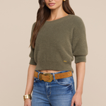 Model wearing an olive green sweater with jeans and a belt on a beige background.