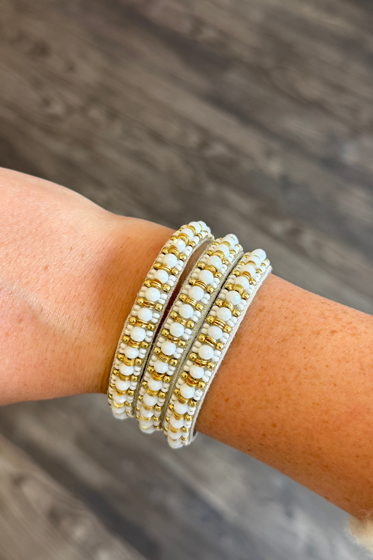 Beaded bracelet with white beads and gold metal links, worn on a person's wrist against a blurred wood background.