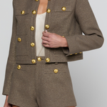 A cropped tweed jacket and matching shorts set in a taupe-brown fabric, featuring prominent gold-tone decorative buttons, modeled on a person against a light gray studio background.