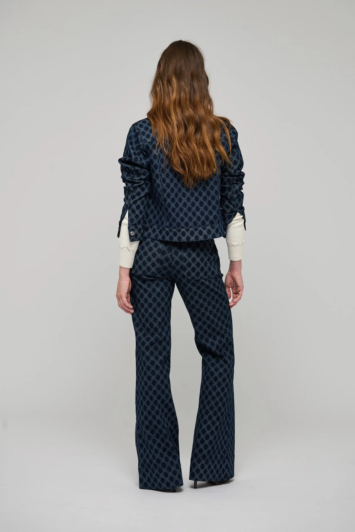 A model viewed from the back wearing a dark navy blue patterned jacket and matching flared pants over a cream long-sleeve shirt, set against a plain light gray studio background.