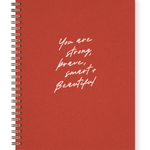 A red spiral-bound notebook with white handwritten-style text on the cover reading 'You are strong, brave, smart, and beautiful', shown on a clean white background.