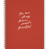 A red spiral-bound notebook with white handwritten-style text on the cover reading 'You are strong, brave, smart, and beautiful', shown on a clean white background.