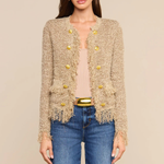 A woman modeling a beige tweed cropped cardigan with gold buttons and fringe trim, styled with a beige top, blue jeans, and a gold belt against a solid beige background.