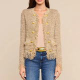 A woman modeling a beige tweed cropped cardigan with gold buttons and fringe trim, styled with a beige top, blue jeans, and a gold belt against a solid beige background.
