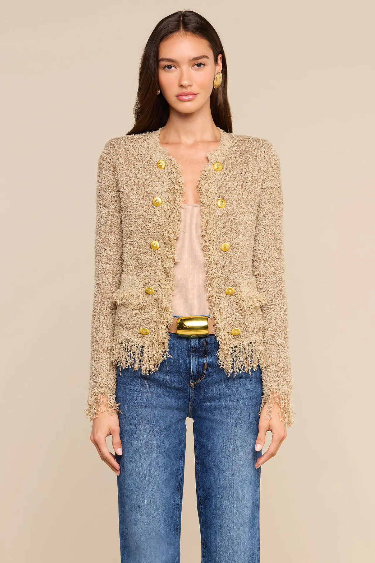 A woman modeling a beige tweed cropped cardigan with gold buttons and fringe trim, styled with a beige top, blue jeans, and a gold belt against a solid beige background.