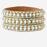 A wide leather cuff bracelet decorated with multiple rows of light blue and white beads accented by small gold beads, shown on a plain light gray background.