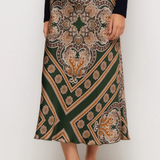 A woman wearing a dark green midi skirt with a detailed beige, brown, and gold paisley and geometric pattern, styled with a black long-sleeve top and black slingback heels against a plain white studio background.