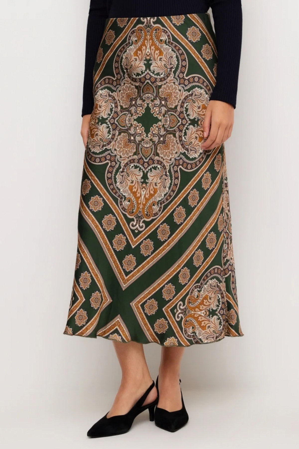 A woman wearing a dark green midi skirt with a detailed beige, brown, and gold paisley and geometric pattern, styled with a black long-sleeve top and black slingback heels against a plain white studio background.