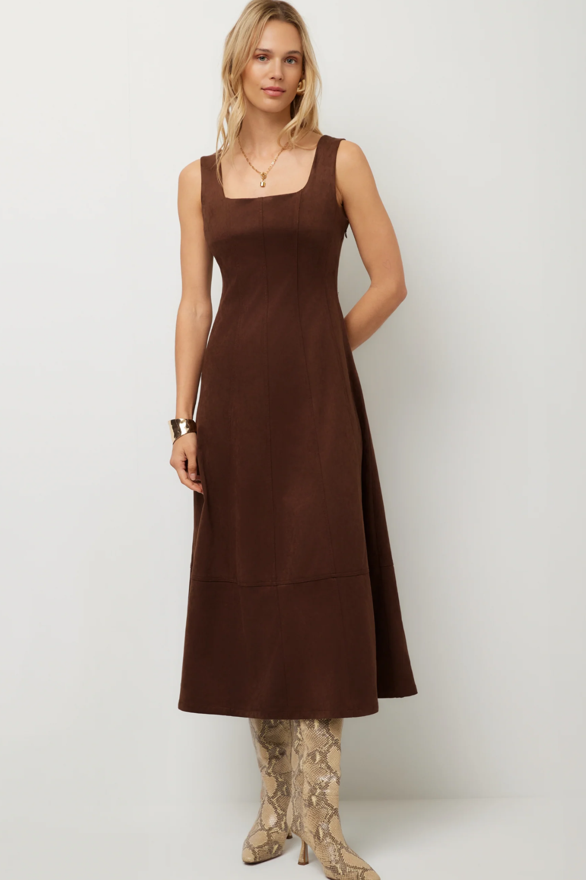 A woman modeling a sleeveless brown midi dress with a square neckline and a flared skirt, paired with snakeskin-patterned boots, against a plain light gray studio background.