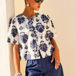 Woman wearing a white blouse with blue floral patterns and blue pants against a beige wall.
