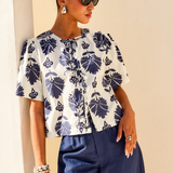 Woman wearing a white blouse with blue floral patterns and blue pants against a beige wall.