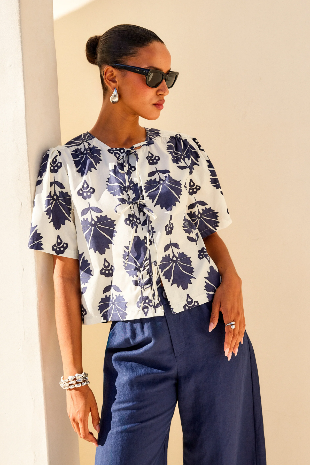 Woman wearing a white blouse with blue floral patterns and blue pants against a beige wall.
