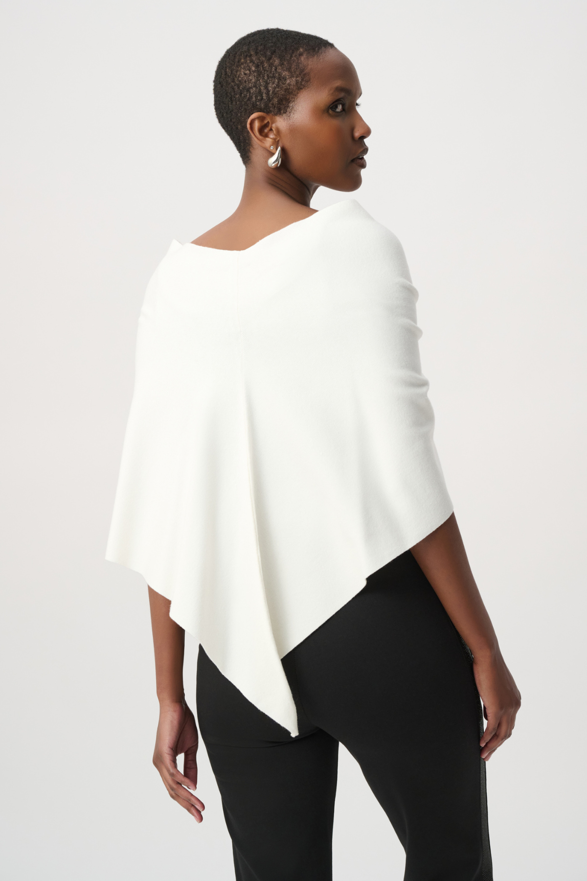 A model wearing a cream-colored wrap shawl with a fluffy pom-pom detail at the center, styled over a black top against a light gray studio background.
