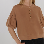 A woman wearing a cropped tan crewneck sweatshirt with puff sleeves and gold-tone decorative buttons down the front, paired with black jeans, against a light gray studio background.