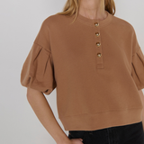 A woman wearing a cropped tan crewneck sweatshirt with puff sleeves and gold-tone decorative buttons down the front, paired with black jeans, against a light gray studio background.