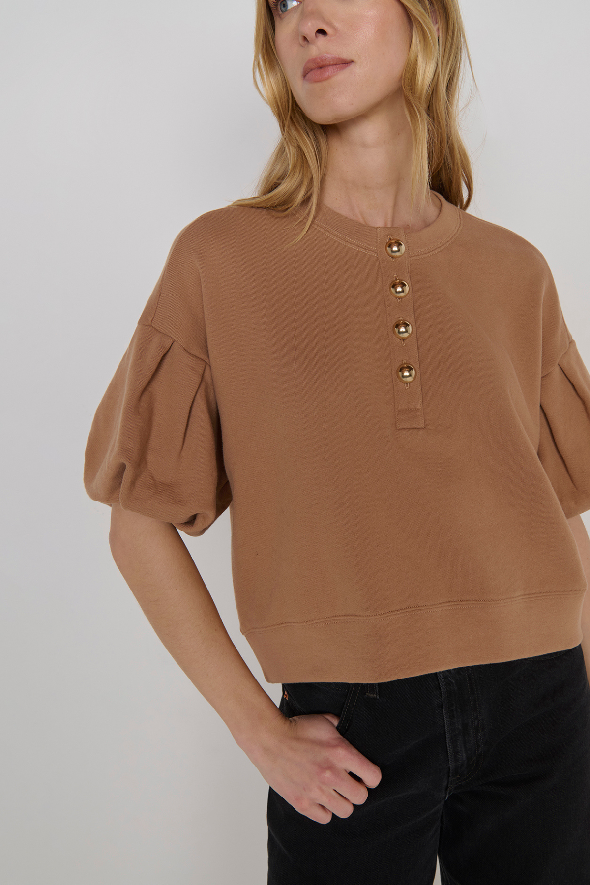 A woman wearing a cropped tan crewneck sweatshirt with puff sleeves and gold-tone decorative buttons down the front, paired with black jeans, against a light gray studio background.