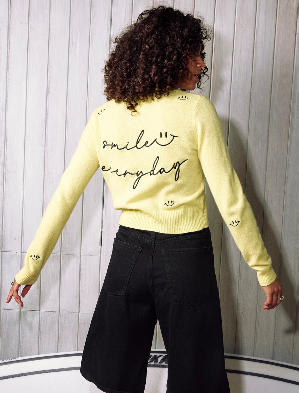 Person wearing a yellow sweater with 'smile everyday' text, standing against a light wooden panel background.