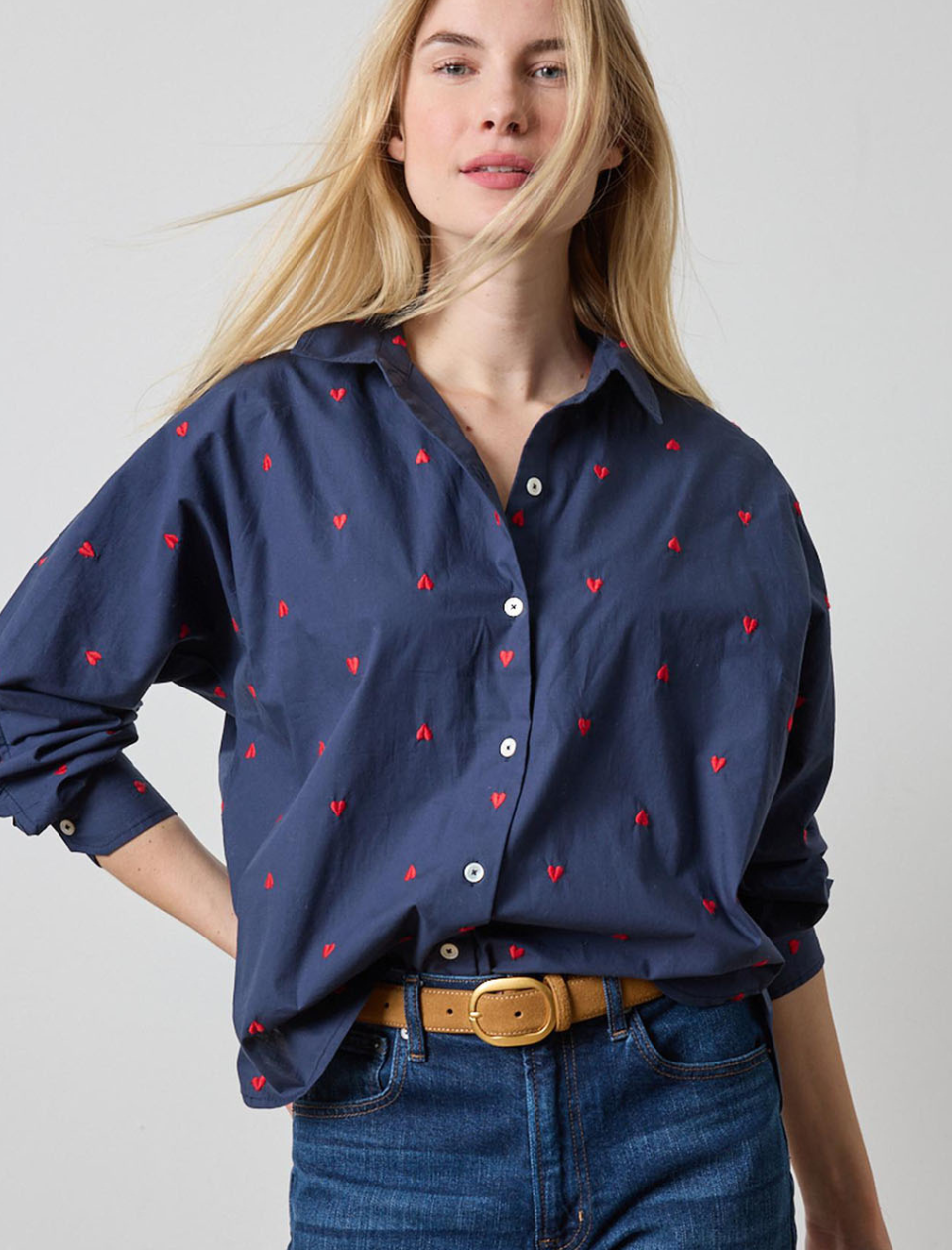 Woman wearing a navy blue shirt with red heart patterns and jeans on a plain background