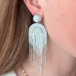 A close-up of a woman's ear wearing a large, dangling statement earring. The earring features a textured silver dome post and a long, fringed arch made of silver beads and chains.