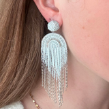 A close-up of a woman's ear wearing a large, dangling statement earring. The earring features a textured silver dome post and a long, fringed arch made of silver beads and chains.