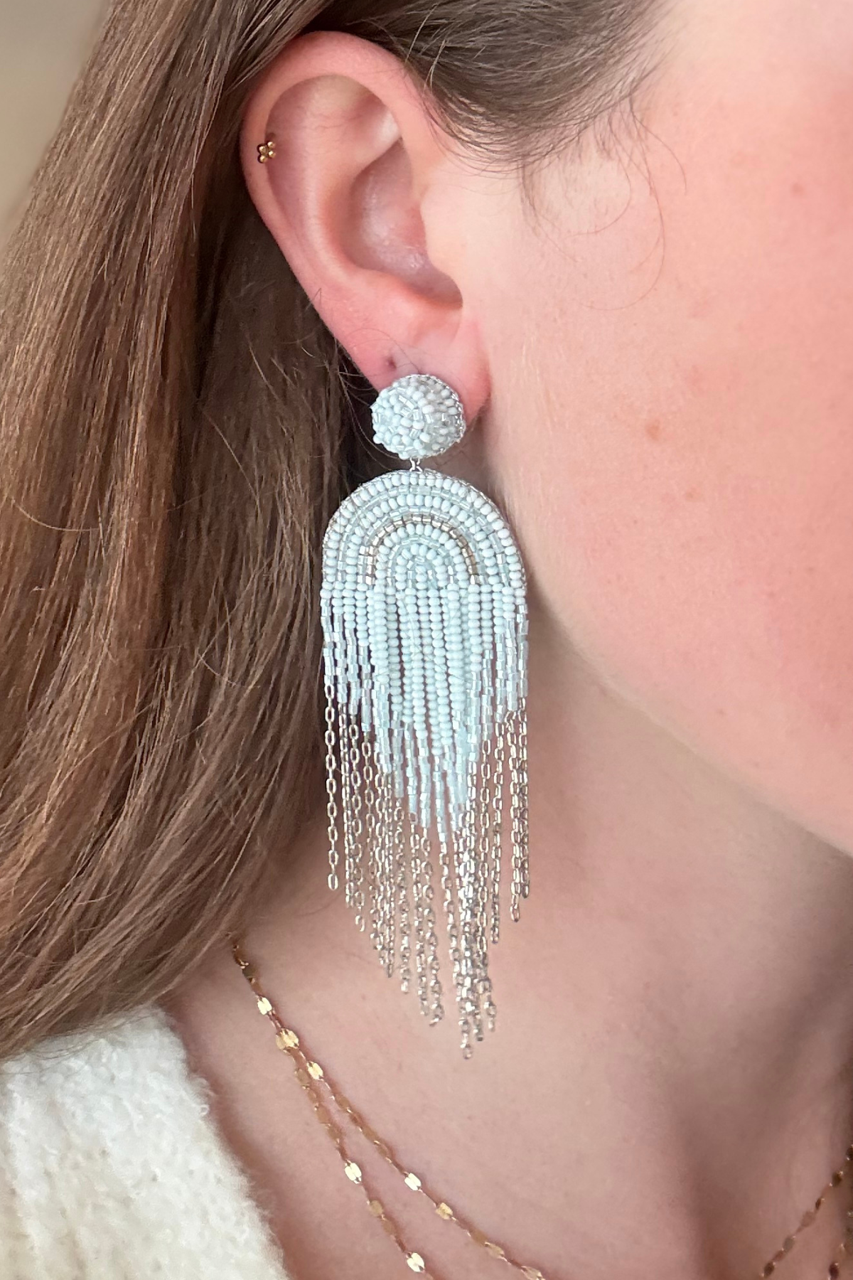 A close-up of a woman's ear wearing a large, dangling statement earring. The earring features a textured silver dome post and a long, fringed arch made of silver beads and chains.