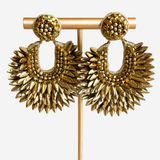 A pair of chunky gold-tone drop earrings displayed on a matching gold stand against a light gray background. The earrings feature textured spherical studs and large, sunburst-shaped dangles with layered beaded and metallic details.